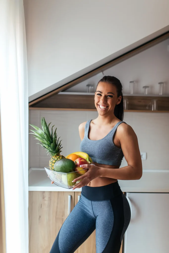 Γυναίκα με αθλητικά ρούχα χαμογελά κρατώντας μπολ με φρέσκα φρούτα στην κουζίνα